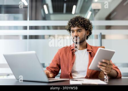 Young man with curly hair focusing on his laptop while holding a digital tablet, working efficiently in a modern office environment, demonstrating pro Stock Photo