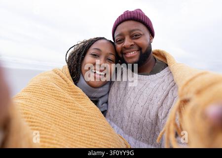 Couple smiling, huddling in mustard-yellow knit blanket on beach, taking smartphone selfie Stock Photo
