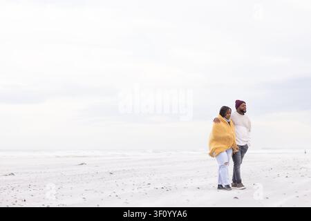 African couple walking with arm draped, wrapped in yellow shawl and beanie on shoreline, copy space Stock Photo
