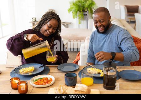 African couple sitting at home dining table, woman pouring juice from pitcher, man cutting toast Stock Photo
