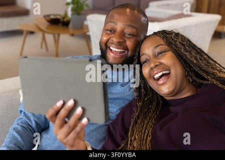 African American couple laughing and sharing gray tablet on sofa in blue knit and plum sweatshirt Stock Photo