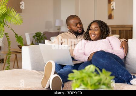 African couple sitting on light sofa, man holding silver laptop, woman wearing pink sweater smiling Stock Photo