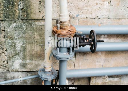 Close-up of an industrial pipeline with a metal valve, rusted flange and PVC pipes mounted on a weathered concrete wall. Concept of water supply, plumbing, utility infrastructure, industrial maintenance, engineering and pipeline systems. Stock Photo