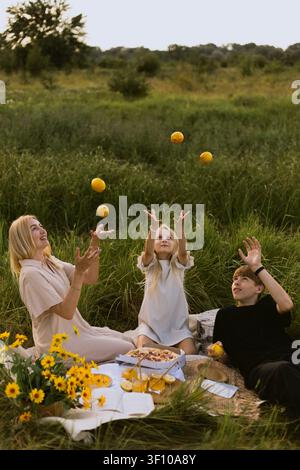 A mother and her two children are having a fun outdoor picnic, tossing lemons in the air amidst tall green grass and wildflowers Stock Photo