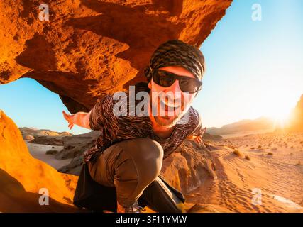 Smiling enthusiastic young man traveler wearing sunglasses headscarf taking action selfie crouching under orange rock arch vast sand dune valley horiz Stock Photo