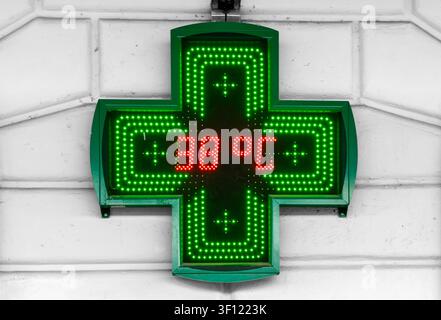 Glowing green LED pharmacy cross sign mounted on a grey wall, showing a hot summer outdoor temperature of 38 degrees Celsius in red digital numbers. Stock Photo