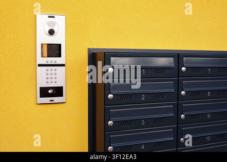 An outdoor multi-apartment IP video intercom system with numerical keypad next to a group of dark grey metal mailboxes mounted on a bright yellow wall Stock Photo