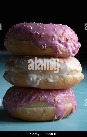 Vibrant tower of sweet ring donuts covered with bright pink and white glaze, decorated with rainbow sprinkles on a blue background Stock Photo