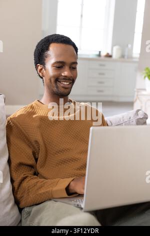 African American man sitting on sofa wearing brown quilted top, using silver laptop at home Stock Photo