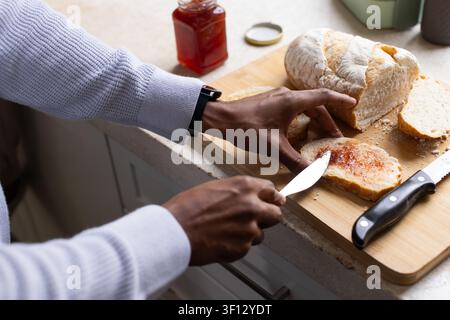 African American man wearing knit sweater and wristwatch spreading jam on bread at kitchen counter Stock Photo
