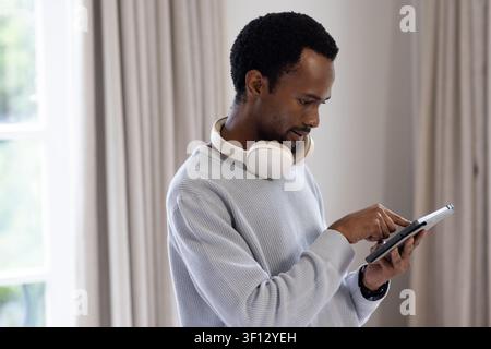 African American man using tablet at home by window, wearing grey sweater, headphones, smartwatch Stock Photo