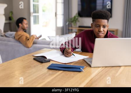Dad and teenage son at home, son studying at table with laptop while dad scrolling smartphone Stock Photo