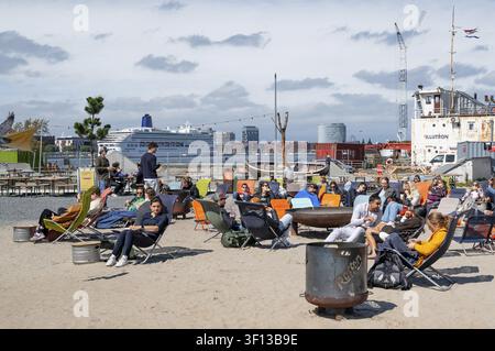 Relaxed people sit in sun loungers on the artificial sandy beach of the Reffen Street Food Market with a view of fire bowls, the Illutron shipwreck an Stock Photo