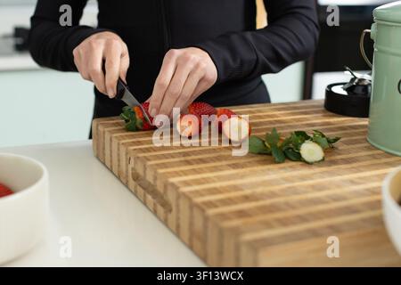 Woman slicing strawberries on butcher block on counter using paring knife in black top in kitchen Stock Photo