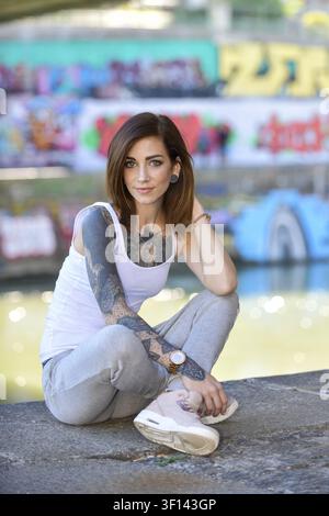 A beautiful young tattooed woman is sitting on the floor next to a river and enjoys the warm summer Stock Photo