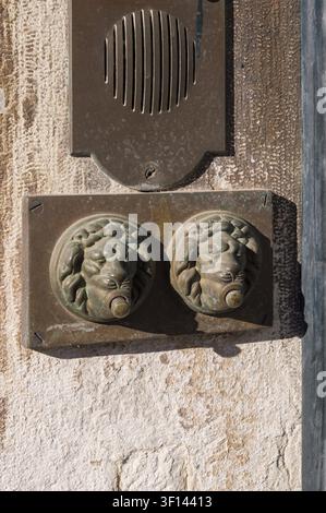 Antique bronze lion-head doorbells mounted on a light beige stone wall. A vintage intercom is also visible Stock Photo