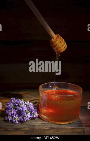 Golden honey pouring from a wooden dipper into a clear glass bowl, with fragrant purple thyme flowers on a rustic wooden surface Stock Photo