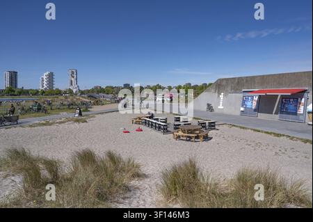 View of a sandy area covered with dune grass in the foreground, on which wooden tables and benches from an outdoor restaurant are placed in front of t Stock Photo