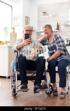Smiling disabled man and his wife using smartphone presents rehabilitation, patient support and assisted recovery with wheelchair, smartphone details in a healthcare or home care setting. Stock Photo