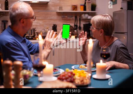 Elderly man and woman wave at a smartphone with green screen while sitting at the kitchen table, using a chroma key mockup for online communication. Stock Photo