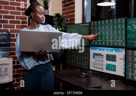 African American stock trader uses a laptop while pointing out high risk investment data on a multi display workstation, showing financial research, trading risk and investment decision context. Stock Photo
