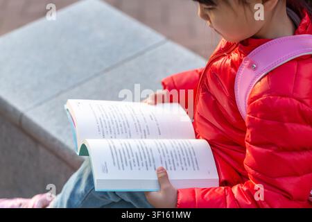 Close up high angle view of a child's hands holding and reading an open book with printed characters, wearing a red down jacket and carrying a pink ba Stock Photo