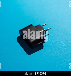 Studio shot of a black USB power adapter with two round European plug pins placed on a bright blue surface, casting a sharp shadow. Stock Photo