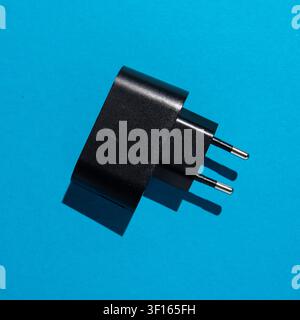 Studio top-view shot of a black wall charger with a two-pin plug placed on a bright blue surface, with strong directional light and a defined shadow. Stock Photo