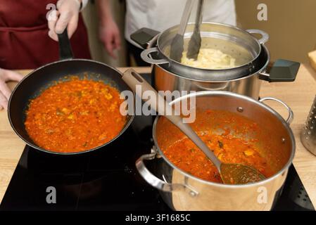 Boiling pasta with tongs in pot and tomato sauce cooking in pans on stove in kitchen for meal cooking. Stock Photo