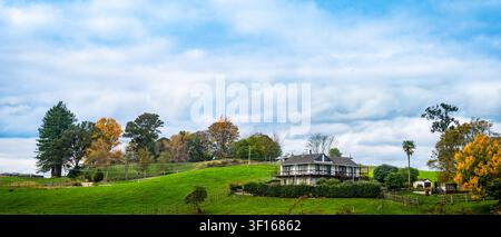 A scenic two-story house nestled on a lush green rolling hillside under a dramatic cloudy sky in New Zealand. Stock Photo