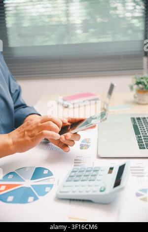 Professional hands using a smartphone beside laptop, calculator, and financial charts on office desk, showing mobile business, accounting review, data Stock Photo