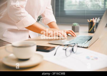 Professional hands typing on laptop at office desk with coffee, smartphone, eyeglasses, and business reports, showing remote work, productivity, digit Stock Photo