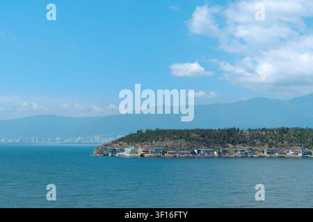 Calm blue sea with a small coastal island, scattered houses, distant mountains and soft clouds under a bright clear sky. Stock Photo