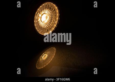 Low-angle view of an illuminated warm white GU10 LED ceiling lamp casting a circular light beam and detailed textured reflection on a dark metallic su Stock Photo