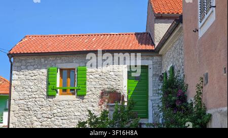 Charming rustic house made of stone with vibrant green wooden shutters and door, red clay tiled roof, flowering plants, and clear blue sky in the back Stock Photo