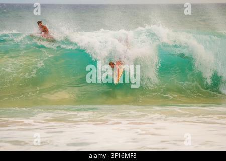 Surfers navigate a powerful breaking turquoise wave near shore, with one duck diving beneath the whitewater as another paddles behind. Stock Photo