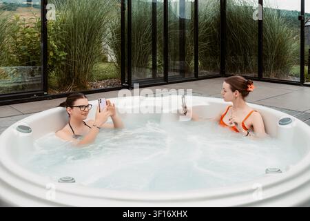 Two women using smartphones while relaxing in an outdoor hot tub at a modern wellness spa retreat Stock Photo