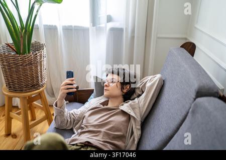 Bored woman relaxing on couch at home using smartphone, browsing social media procrastinating time. Stock Photo