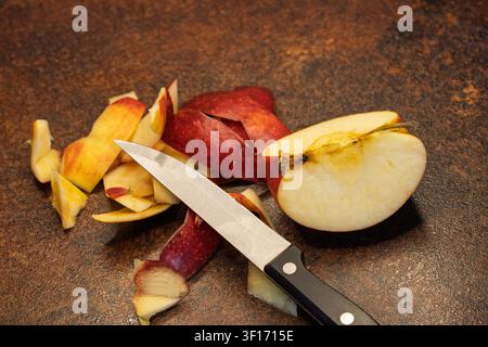 Studio close-up shot of a single fresh red apple slice, a sharp kitchen knife, and scattered apple peels, arranged on a dark, rustic, textured kitchen Stock Photo
