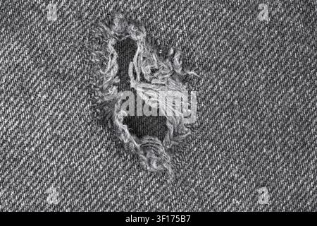 Top view of distressed denim with frayed hole revealing cotton threads. Monochrome texture highlights weave pattern wear. Ideal for fashion, textile, Stock Photo