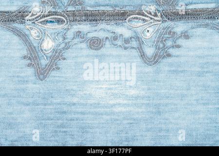 Close up of light blue denim with ornate white embroidery visible stitching. Textured fabric surface, soft lighting highlights weave thread details Stock Photo