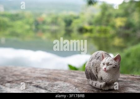 Chiang Mai, Thailand - 19 June 2026 : A grey tabby cat figurine curled up sleeping on a wooden log with copy space, lake and forest blurred in the bac Stock Photo