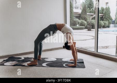 Woman practicing a full wheel pose on a yoga mat in a bright home studio, advanced backbend for spinal mobility, functional strength, body awareness Stock Photo