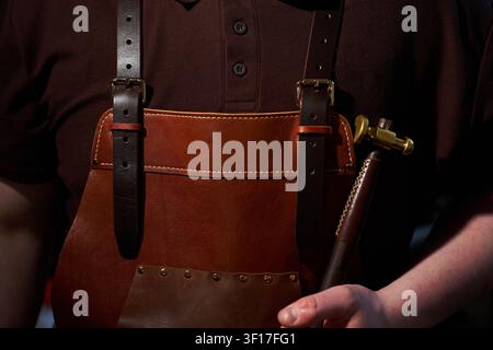 Leatherworker in a handcrafted brown apron holding a specialized brass hammer close up Stock Photo