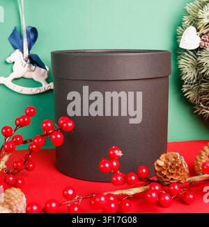 Christmas mockup black gift box on red table against green background. New year cylindrical present package with winter berries pine cones for wreath Stock Photo