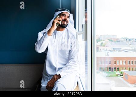 Young arab man wearing traditional emirati kandura and ghutra, smiling while calling and looking out a window, representing business communication and Stock Photo