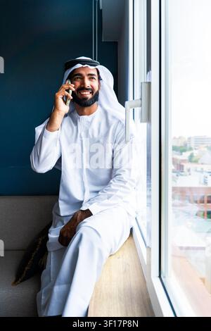 Arab man wearing traditional emirati kandura and ghutra, smiling while having a phone conversation, looking away, sitting by a bright window, showcasi Stock Photo