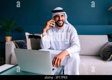 Arab man wearing a kandura and ghutra sitting on a sofa, happily talking on a smartphone while using a laptop, representing remote work and modern bus Stock Photo