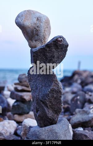 Balanced stones are stacked on a seashore, showcasing unique shapes and textures, with the ocean and sunset visible in the background Stock Photo