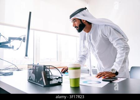 Arab businessman concentrating on a computer screen while leaning over an office desk with paperwork, coffee cup, and smartphone, representing modern Stock Photo